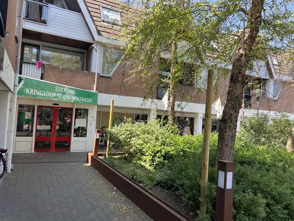 Gevel van kringloopwinkel met groene letters, rode deuren en houten plantenbakken ervoor