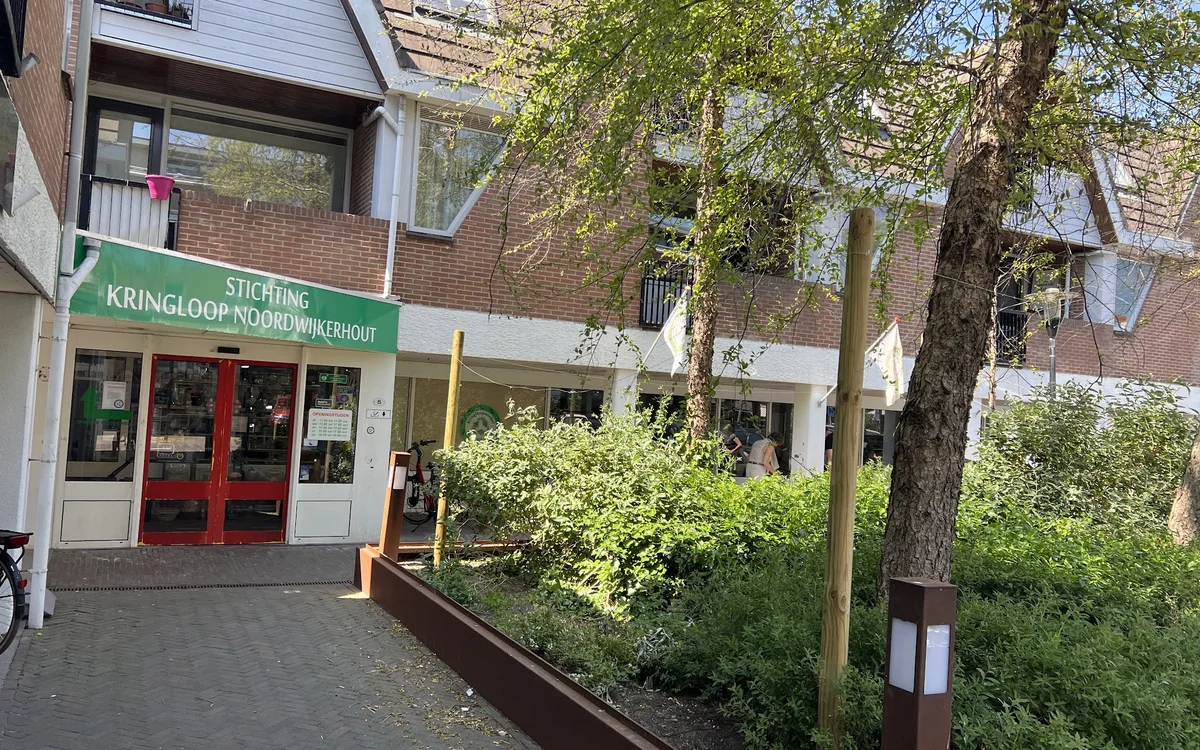 Gevel van kringloopwinkel met groene letters, rode deuren en houten plantenbakken ervoor