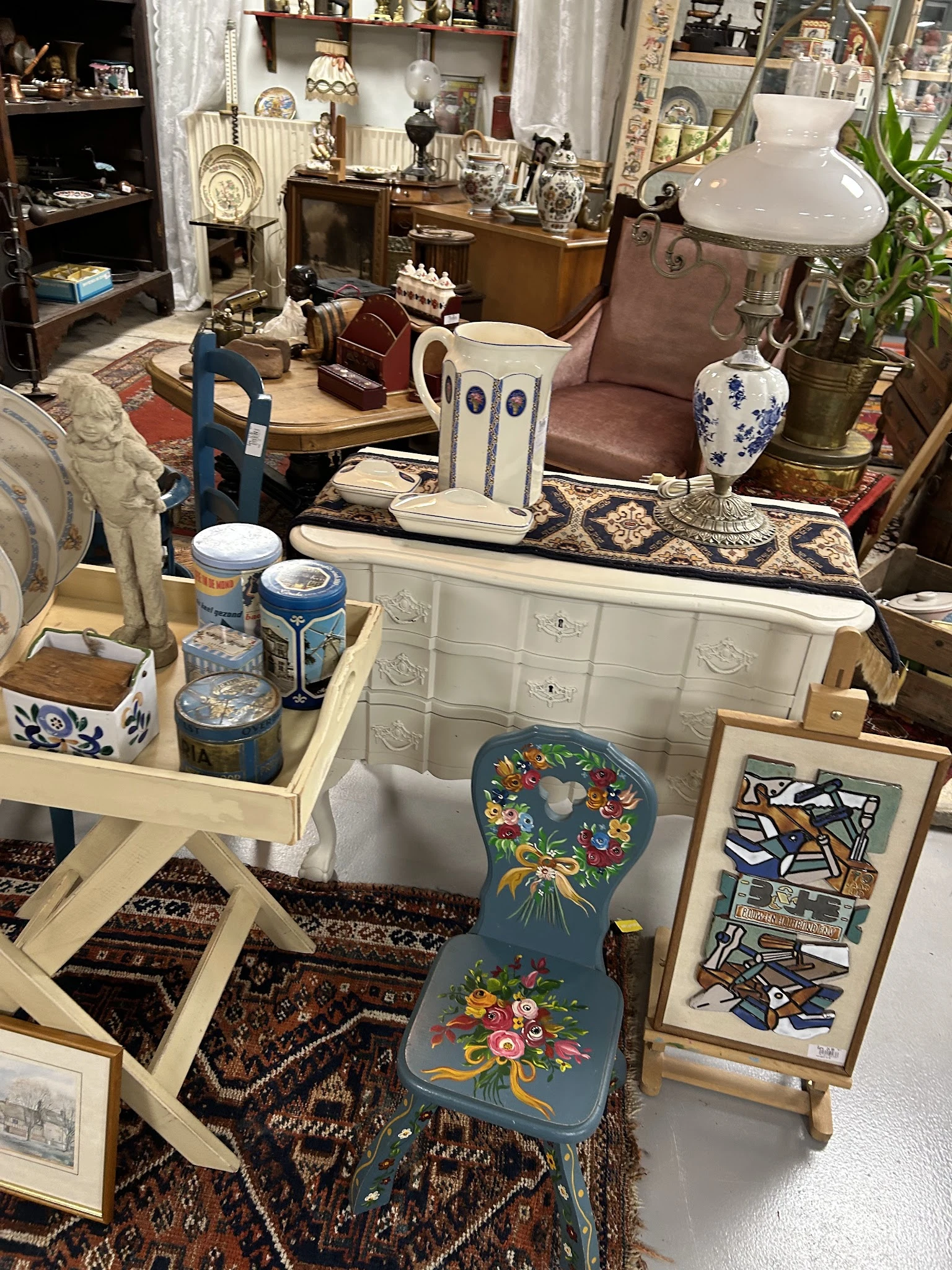 Uitgestalde tweedehands meubels en decoratiestukken inclusief witte kast, blauwe kruk, aardewerk en kunstobjecten