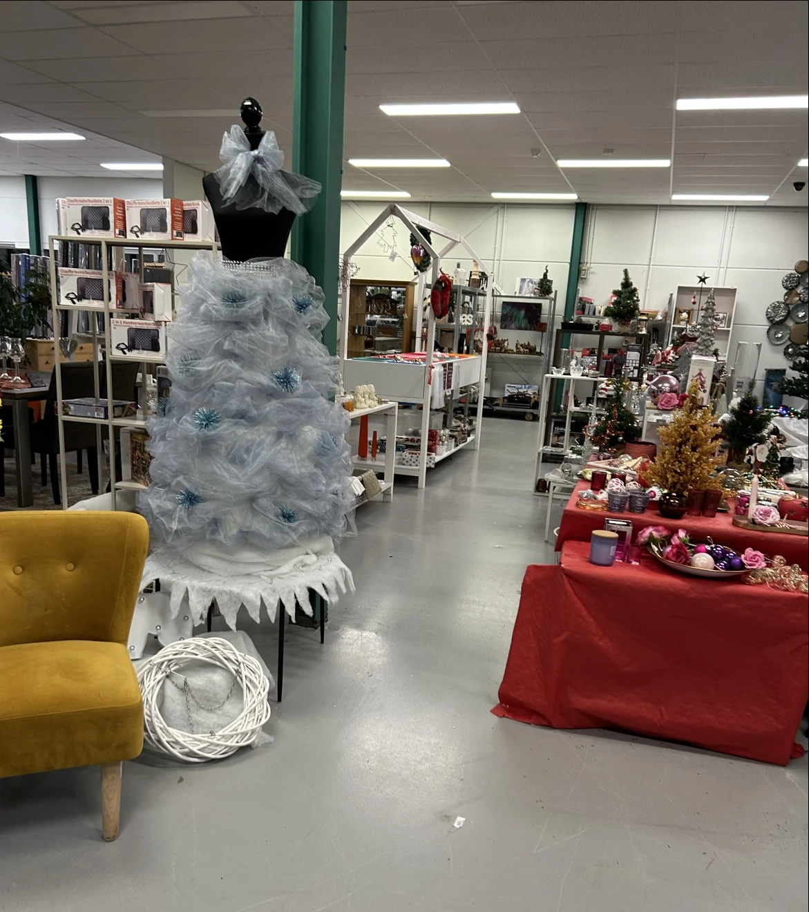 Winkelvloer met kerstdecoratie, blauwe kunstkerstboom, rode tafels met kerstspullen en gele fauteuil