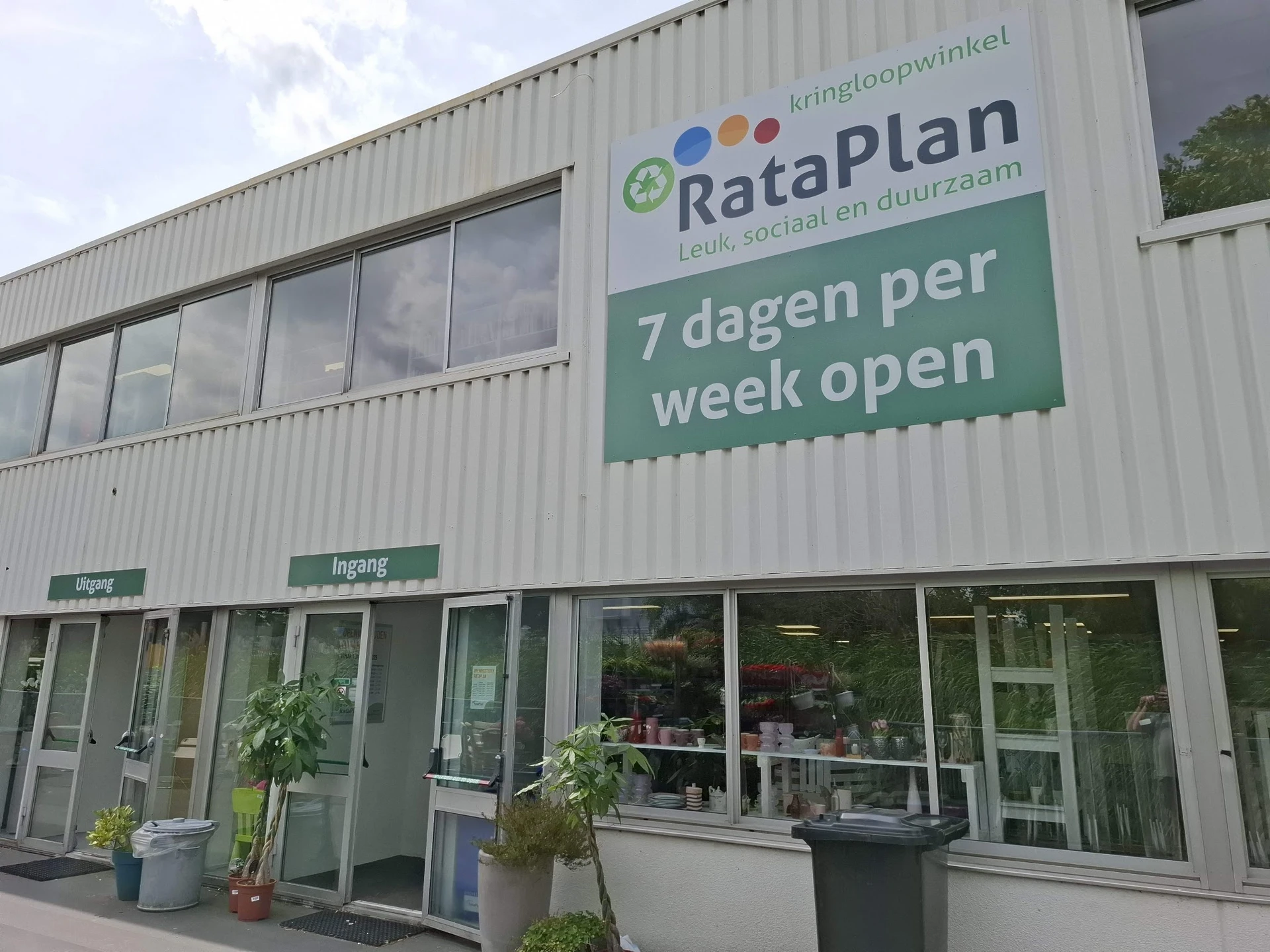 Voorgevel van kringloopwinkel RataPlan met groene en witte signage, twee ingangsdeuren en ramen, met informatie '7 dagen per 