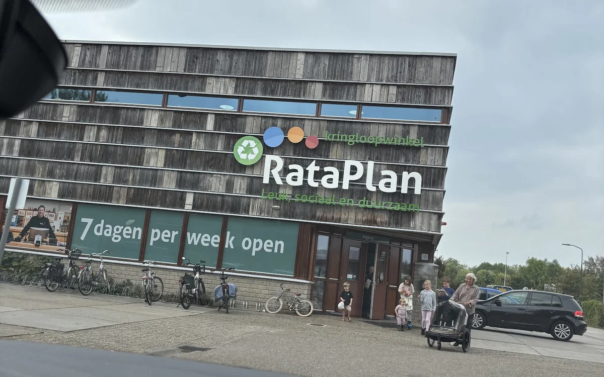 Gevel van RataPlan kringloopwinkel met groene banner, fietsen ervoor geparkeerd en bezoekers op het plein