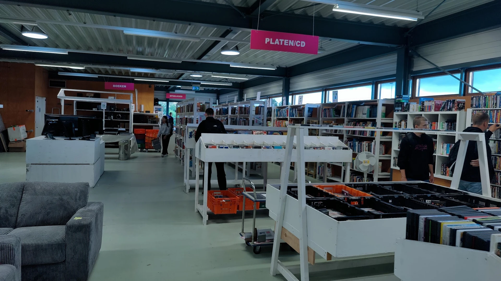 Overzicht van kringloopwinkel met witte stellingen, boekenrekken, diverse goederen en winkelgang met plafondbordje 'PLATENICD