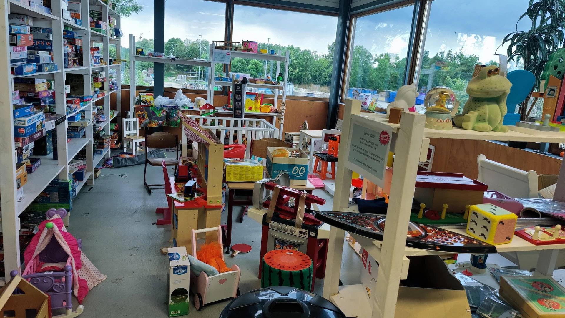 Speelgoedafdeling in kringloopwinkel met kinderspeelgoed, poppen, puzzels en speelhuisjes bij grote ramen met uitzicht op gro