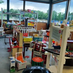 Speelgoedafdeling in kringloopwinkel met kinderspeelgoed, poppen, puzzels en speelhuisjes bij grote ramen met uitzicht op gro
