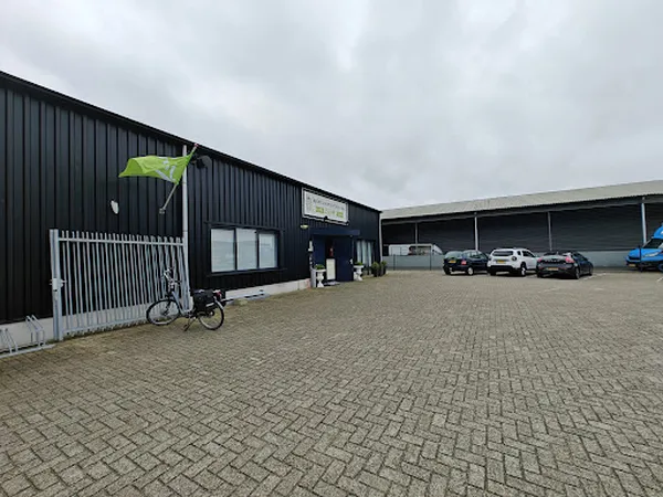 Voorgevel van donkere loods met witte deur, ingang en naam op pui, parkeerplaats met enkele auto's