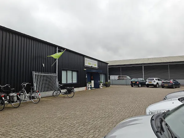 Voorgevel van kringloopwinkel met zwarte golfplaten wand, ingang, parkeerplaats met auto's en fietsen