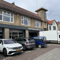 Gevel van kringloopwinkel met bakstenen voorgevel, grote ramen, en parkeerplaats met auto's