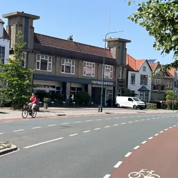 Straatgevel van winkel aan Geversstraat met bruin pand, grote ramen, twee schoorstenen en kantelpui op begane grond