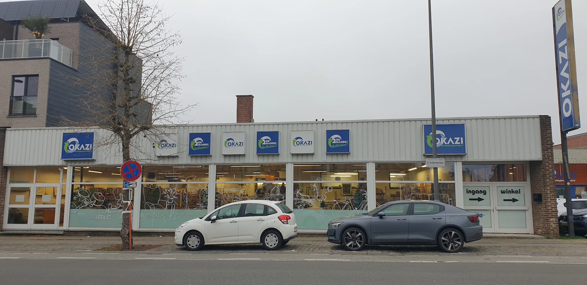 Gevel van Okazi kringloopwinkel met blauwe logos op grijze golfplaten, twee auto's geparkeerd ervoor
