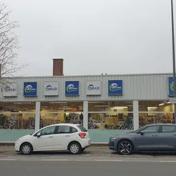 Gevel van Okazi kringloopwinkel met blauwe logos op grijze golfplaten, twee auto's geparkeerd ervoor