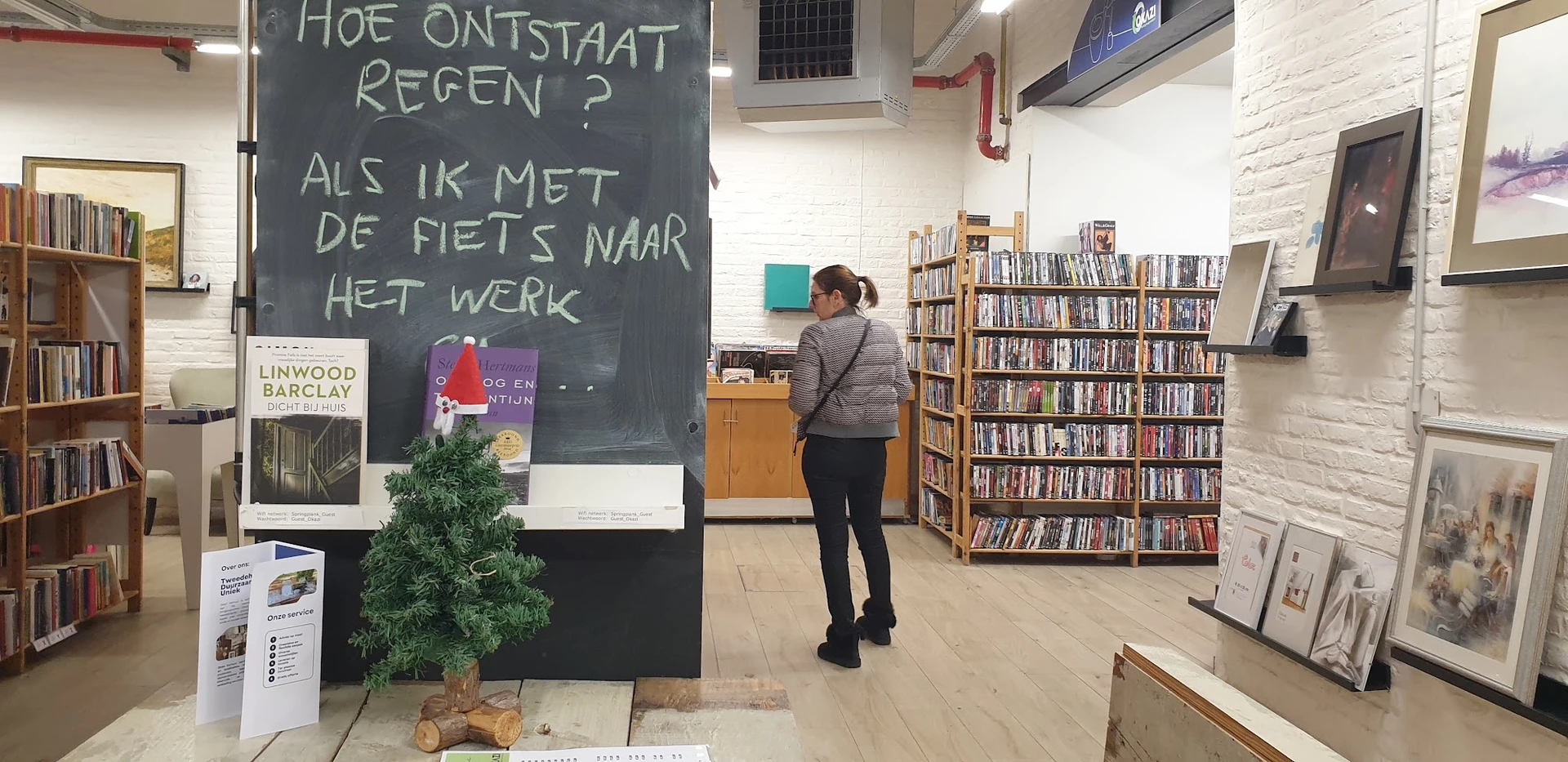 Interieur van kringloopwinkel met boekenrekken, krijtbord met Nederlands tekstbord en bezoekers die rondkijken