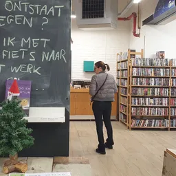 Interieur van kringloopwinkel met boekenrekken, krijtbord met Nederlands tekstbord en bezoekers die rondkijken