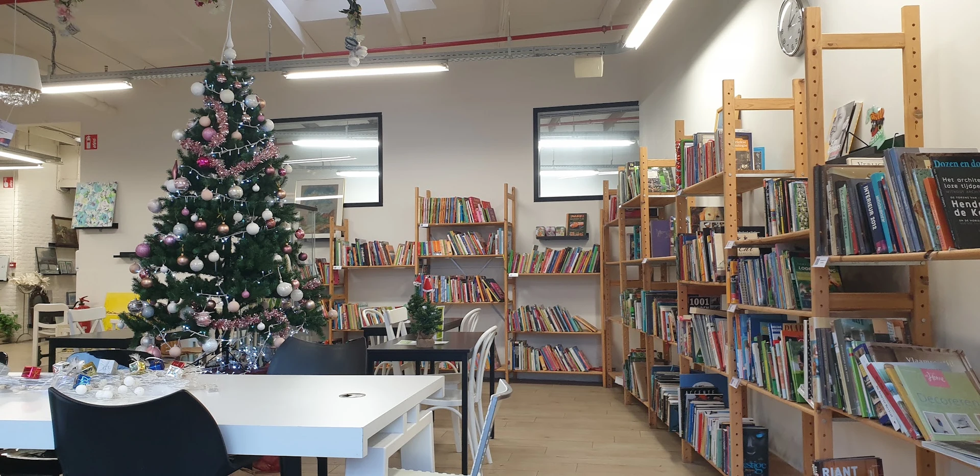 Binnenaanzicht van een kringloopwinkel met houten boekenkasten, een versierde kerstboom en leeshoek met tafel en stoelen