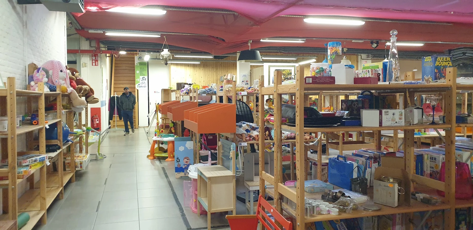 Winkelvloer van kringloopwinkel met houten stellingen vol spullen, toonbank en klanten in het achtergrond