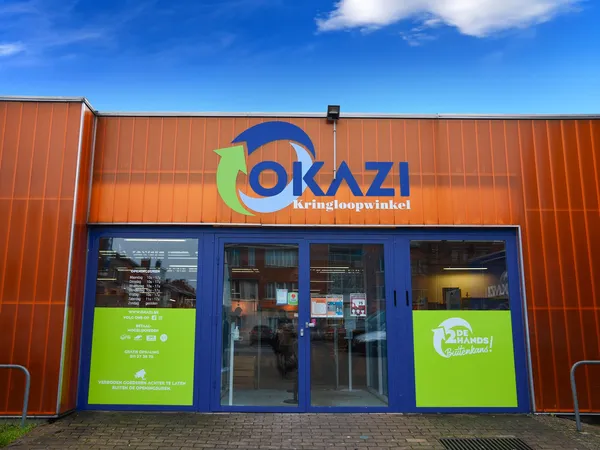 Voorkant van kringloopwinkel Okazi met oranje golfplaten gevel, blauwe ramen en groen logo