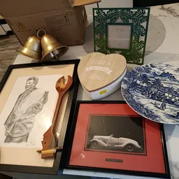 Diverse tweedehands items op marmeren tafel: ingelijste kunstprenten, decoratieve borden, houten bestek, gouden bellen en fot