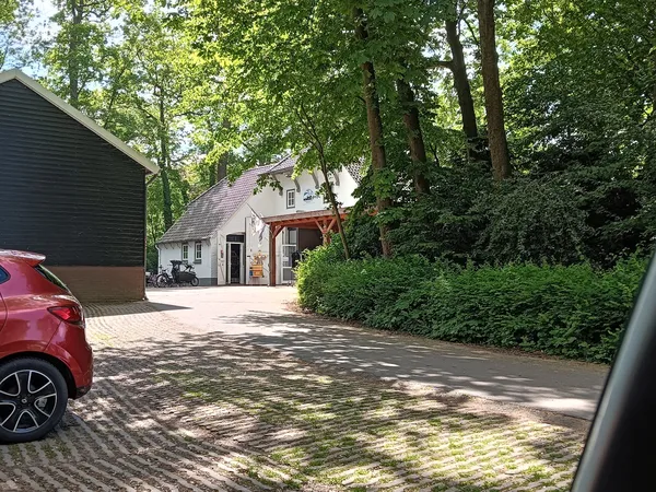 Voorkant van kringloopwinkel met witte gevel, houten overkapping en parkeerplaats met bakstenen bestrating