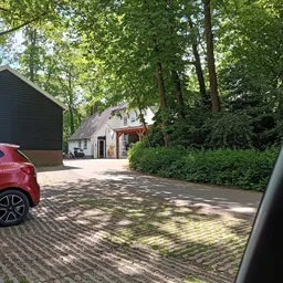 Voorkant van kringloopwinkel met witte gevel, houten overkapping en parkeerplaats met bakstenen bestrating