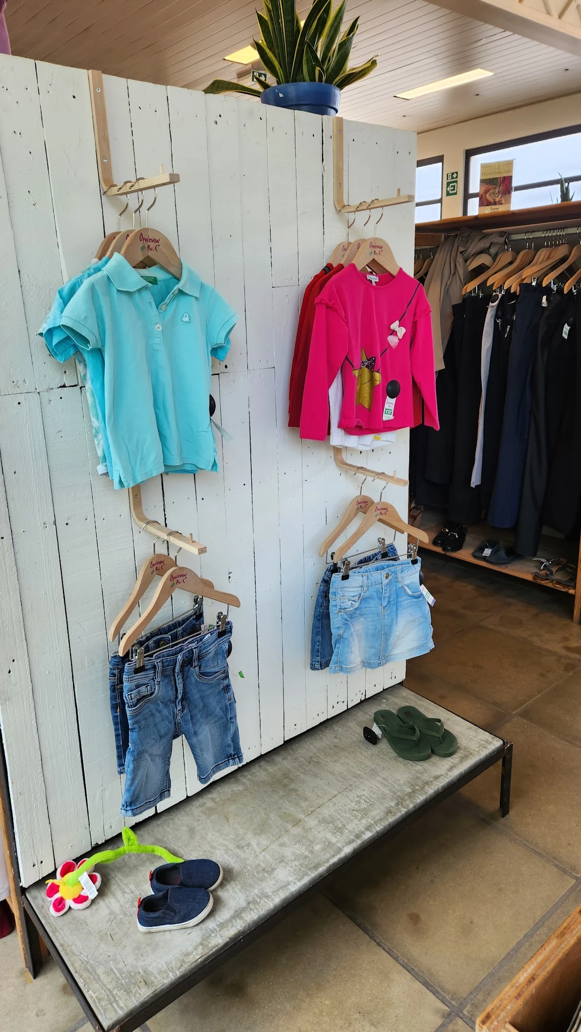 Tweedehands kleding uitgestald op houten hangers aan witte wand: blauwe polo, roze vest, zwarte blazers en spijkerbroeken