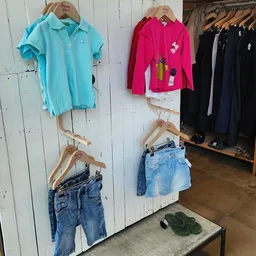 Tweedehands kleding uitgestald op houten hangers aan witte wand: blauwe polo, roze vest, zwarte blazers en spijkerbroeken