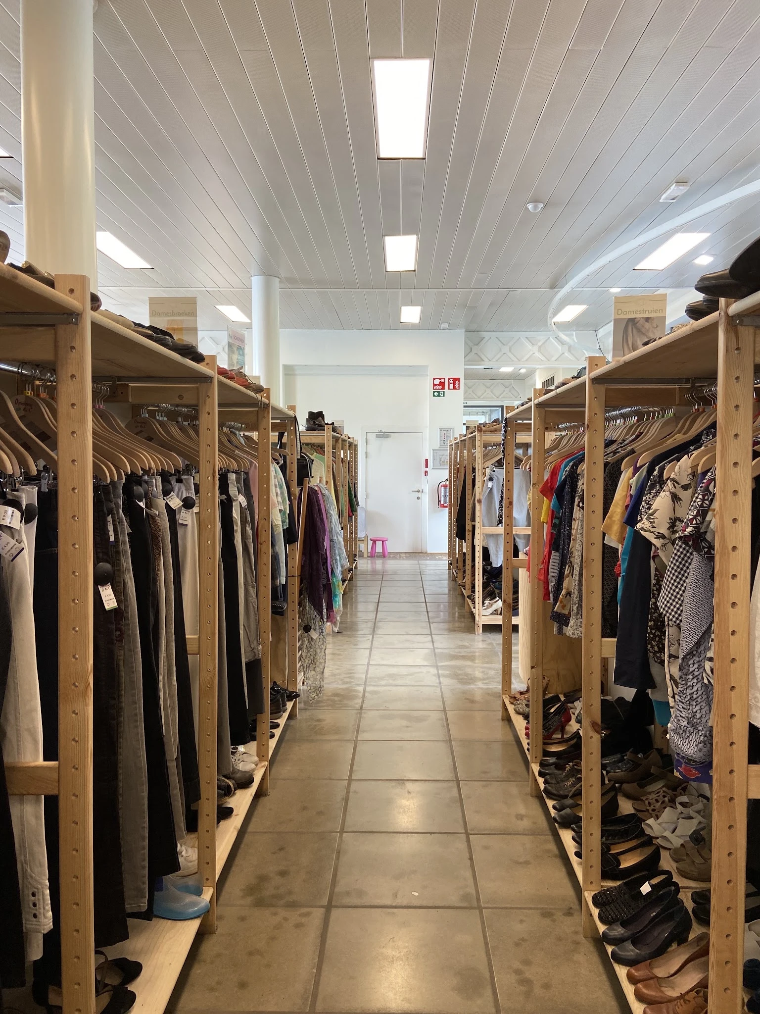 Lange gang met houten stellingen vol tweedehands kleding aan beide zijden, goed verlicht