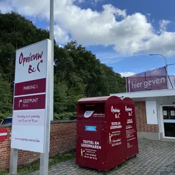 Voorgevel van kringloopwinkel met witte zijgevel, rode infokasten en parkeerinstructies, omgeven door groen.