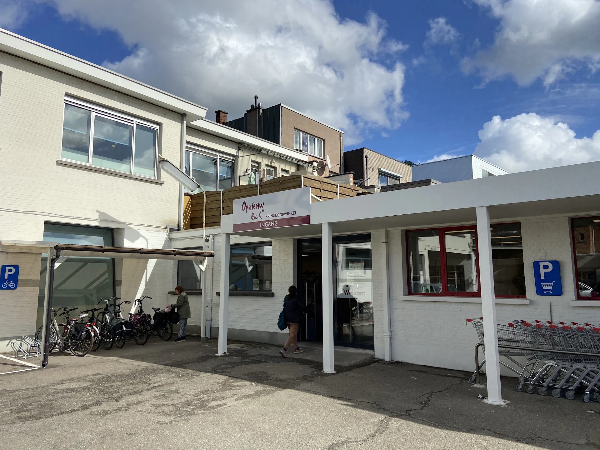 Winkelvoorgevel met witte afdakking, glazen deuren en ramen, fietsenstalling aan zijkant, parkeerplaatsen en winkelwagentjes