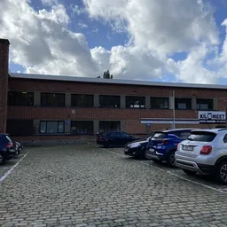 Voorkant van een bakstenen kringloopwinkel met parkeerplaats en enkele geparkeerde auto's