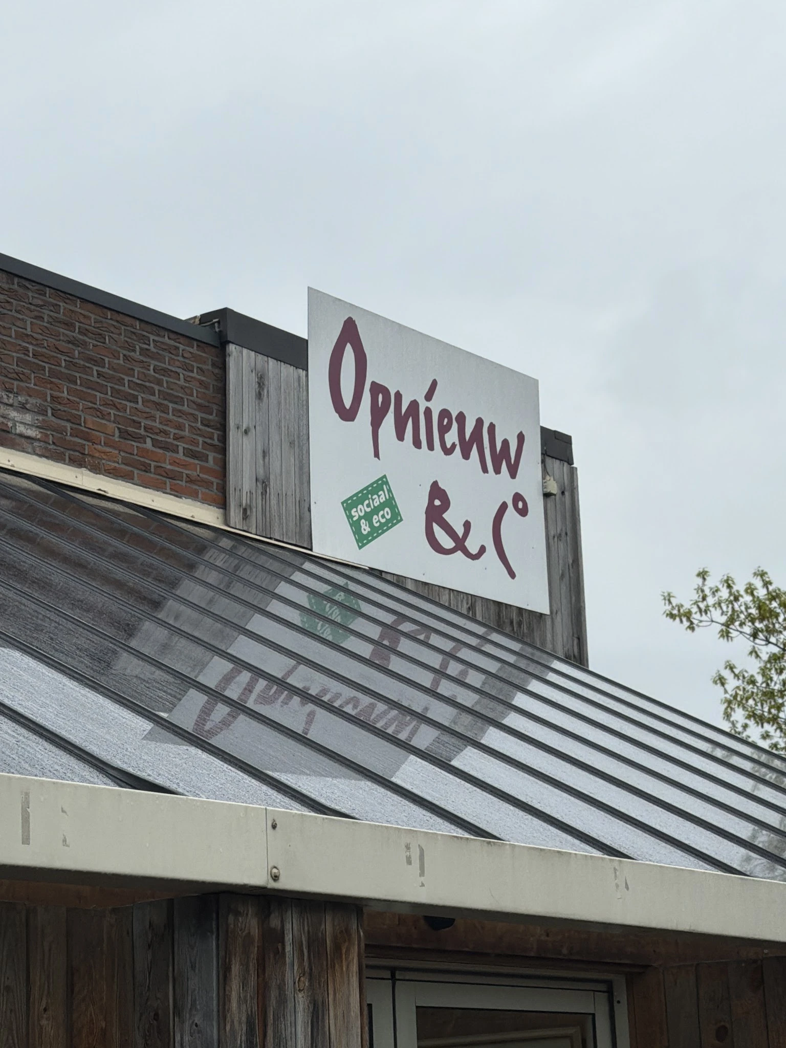Uithangbord van Opnieuw & Co op een dakrand met bakstenen gevel ernaast