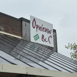 Uithangbord van Opnieuw & Co op een dakrand met bakstenen gevel ernaast