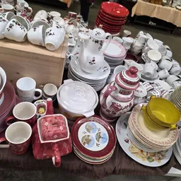 Uitgestalde serviesgoed en keukengerei op een tafel: borden, kopjes, schalen en ketels in wit, rood en roze