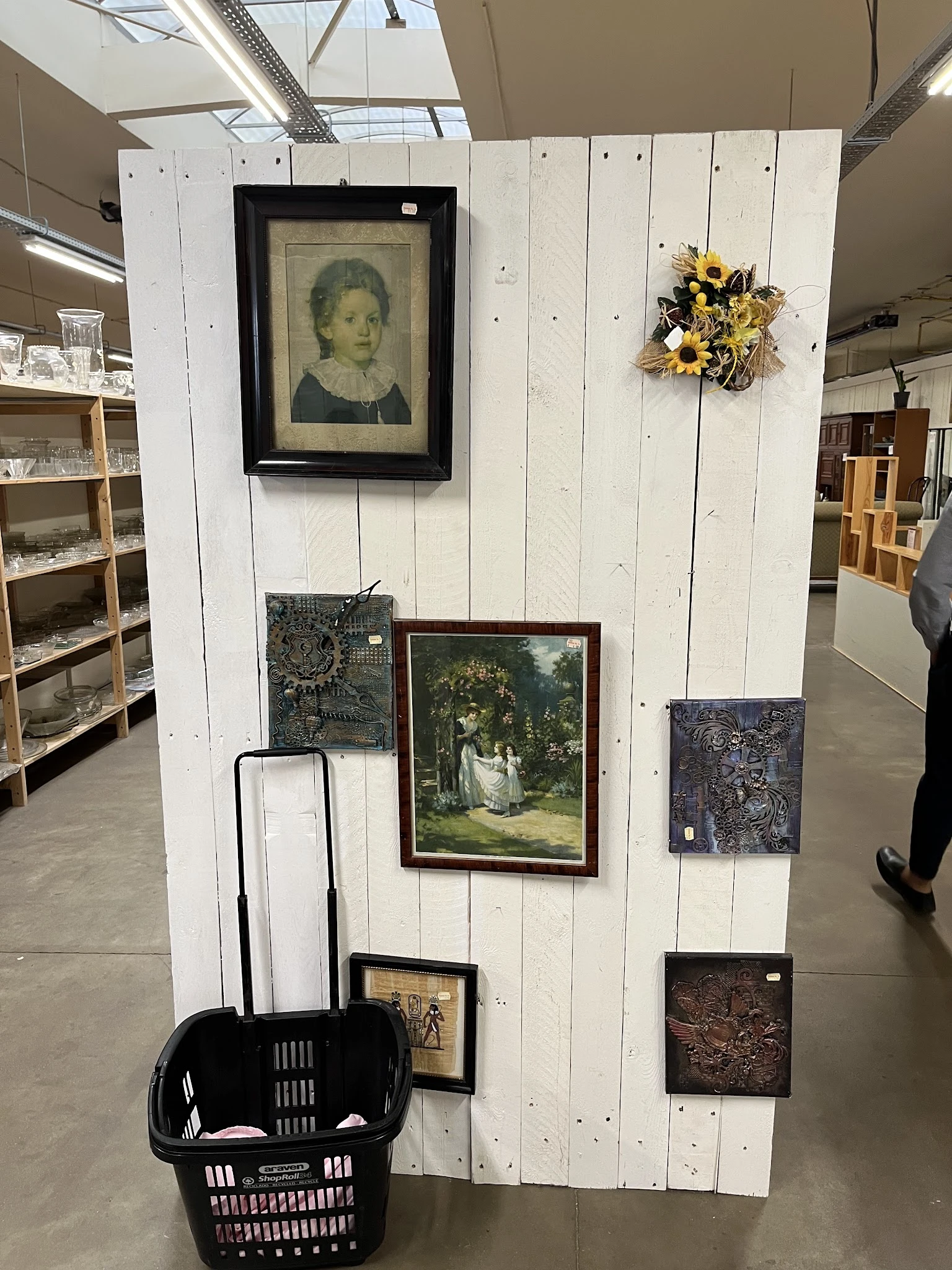 Witte wand met ingelijste kunstwerken, een bloemstuk en houten rekken met spullen in kringloopwinkel
