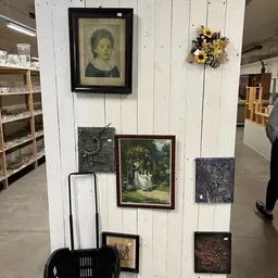 Witte wand met ingelijste kunstwerken, een bloemstuk en houten rekken met spullen in kringloopwinkel