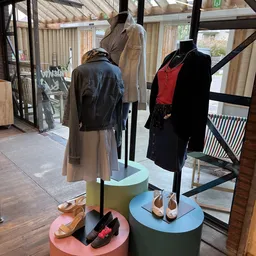 Etalage met kledingmanequins: zwart leren jack met beige plissérok, zwart wollen jas met rood topje, en bruine schoenen op di