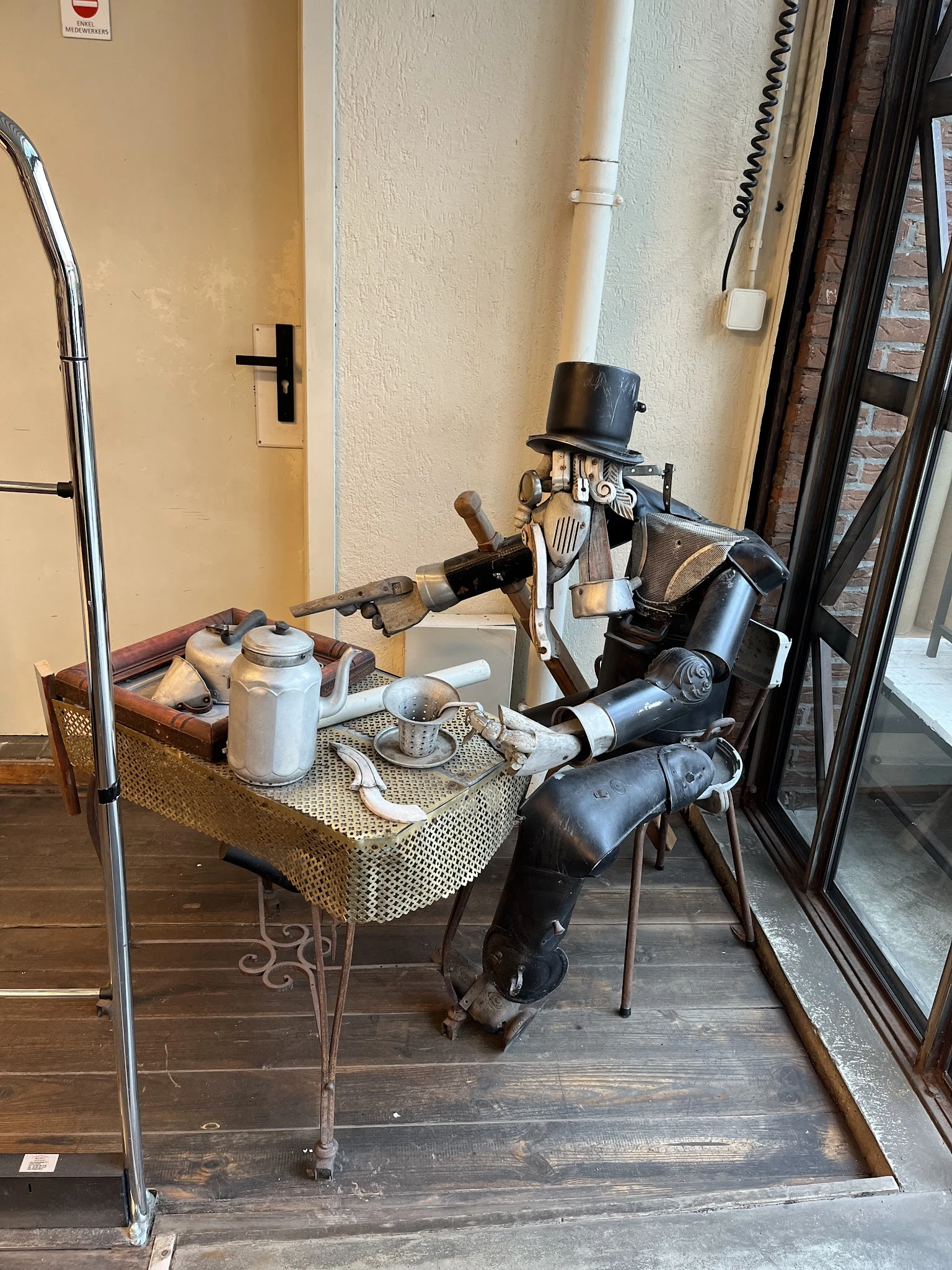 Decoratieve robot-figuur in zwart metaal met hoed zittend aan een tafeltje met vintage huisraad