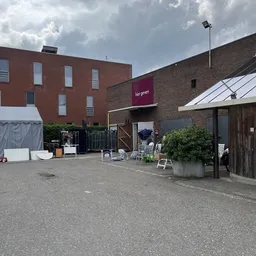 Voorkant van kringloopwinkel met bakstenen gevel, terras met stoelen en witte tent links