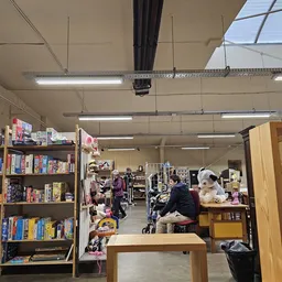 Overzicht van de winkelvloer met boekenrekken aan de linkerkant, kleding en diverse spullen in het midden, en houten kastjes 