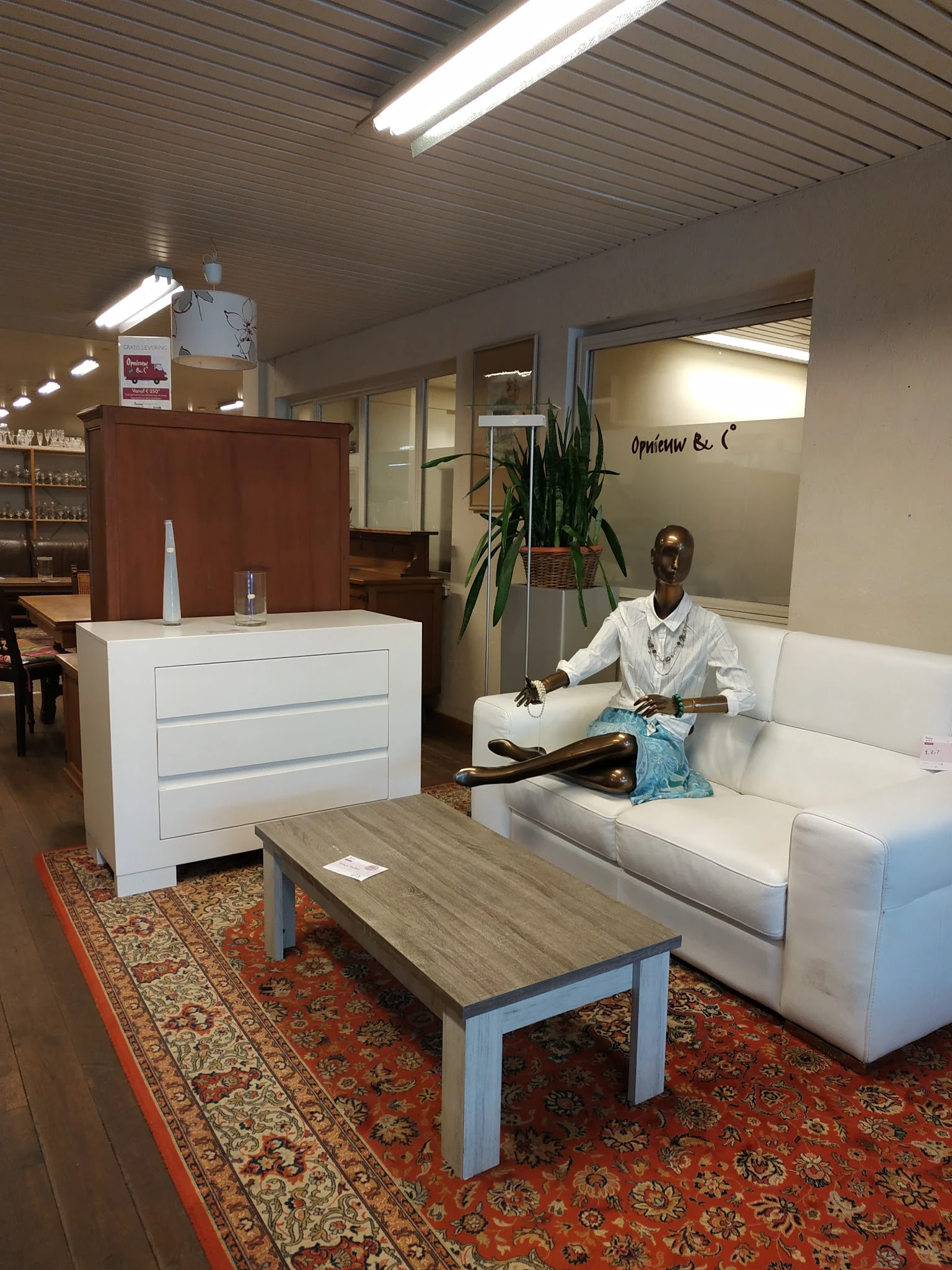 Showroom van kringloopwinkel met witte kast, houten tafel, witte bank en pop-upfiguur, plant en winkelinrichting zichtbaar