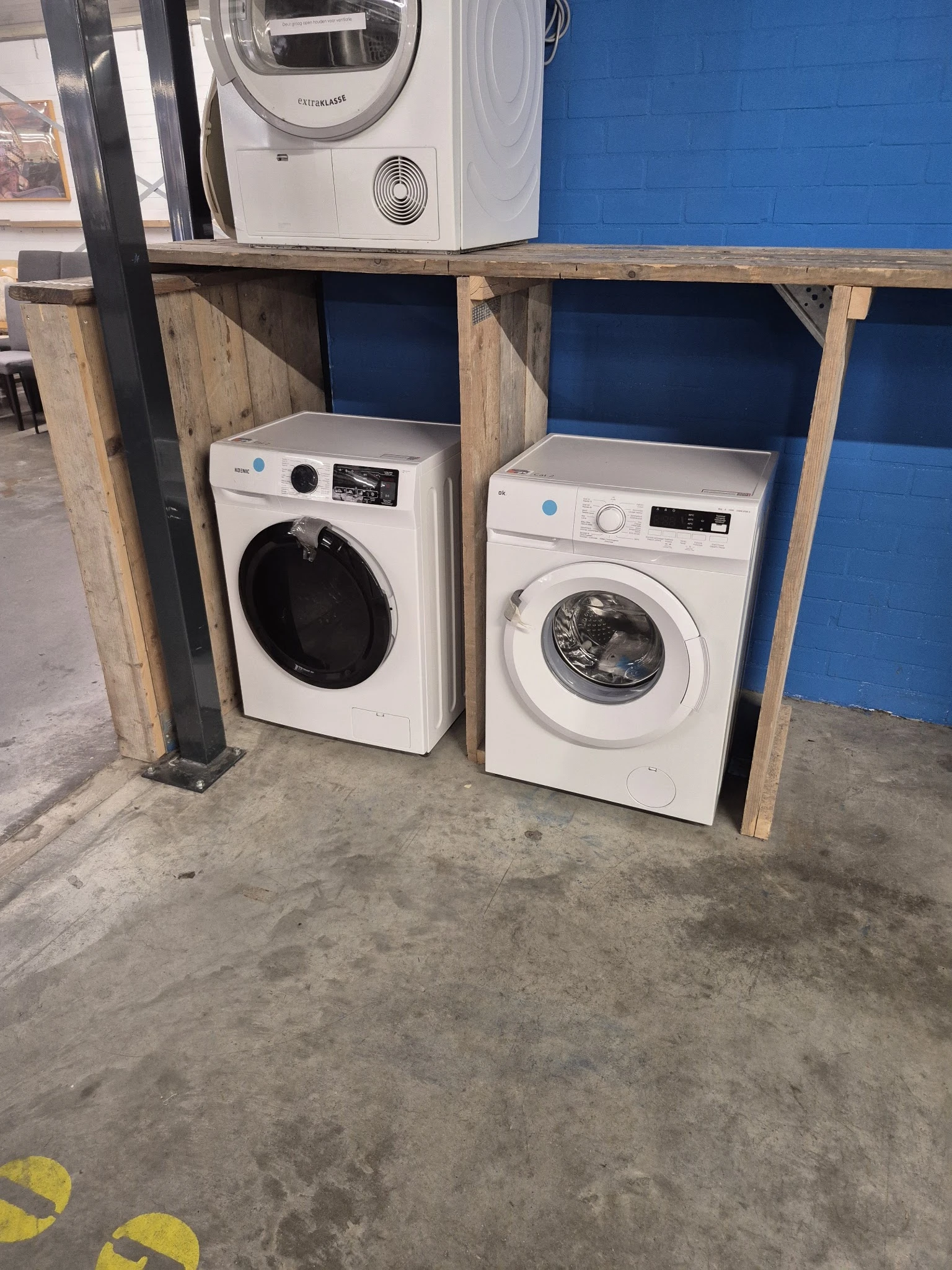 Twee witte wasmachines in een kringloopwinkel, onder houten afdak met blauwe achtergrond