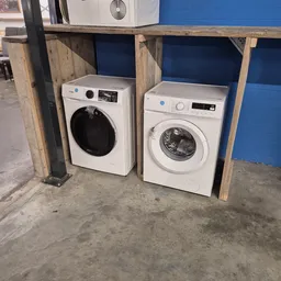 Twee witte wasmachines in een kringloopwinkel, onder houten afdak met blauwe achtergrond