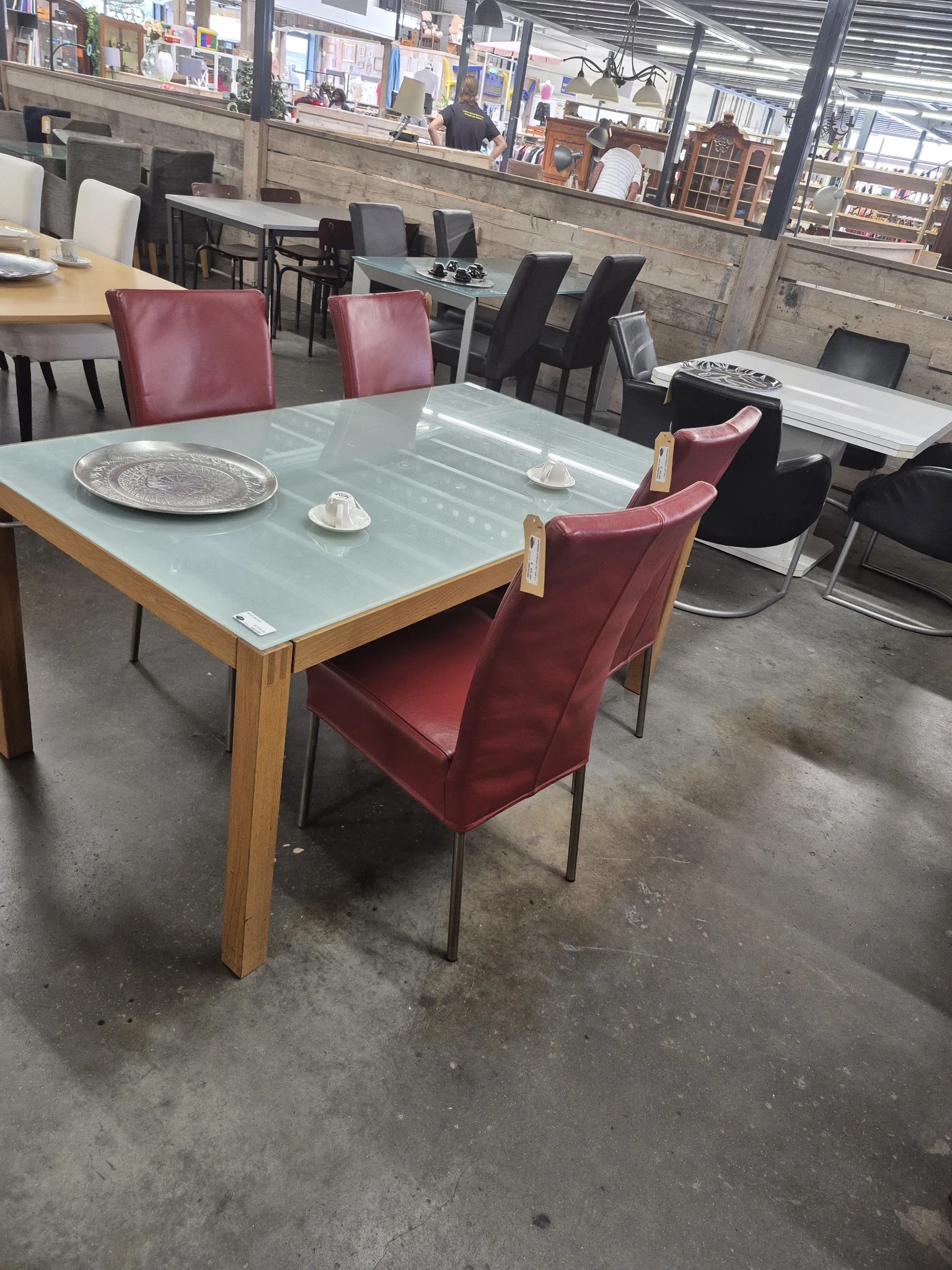 Eetkamertafel met glazen blad en houten onderstel, omringd door bordeauxrode fluwelen stoelen in een kringloopwinkel.