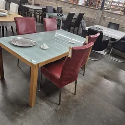 Eetkamertafel met glazen blad en houten onderstel, omringd door bordeauxrode fluwelen stoelen in een kringloopwinkel.
