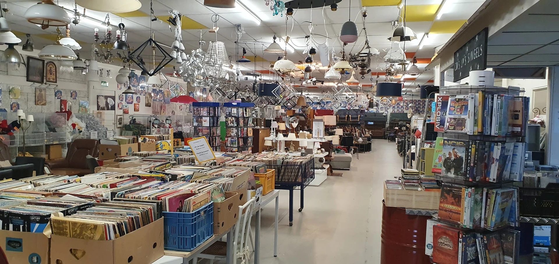 Winkelvloer van kringloopwinkel met diverse hanglampen, stellingen met boeken, meubels en spullen verspreid over de ruimte