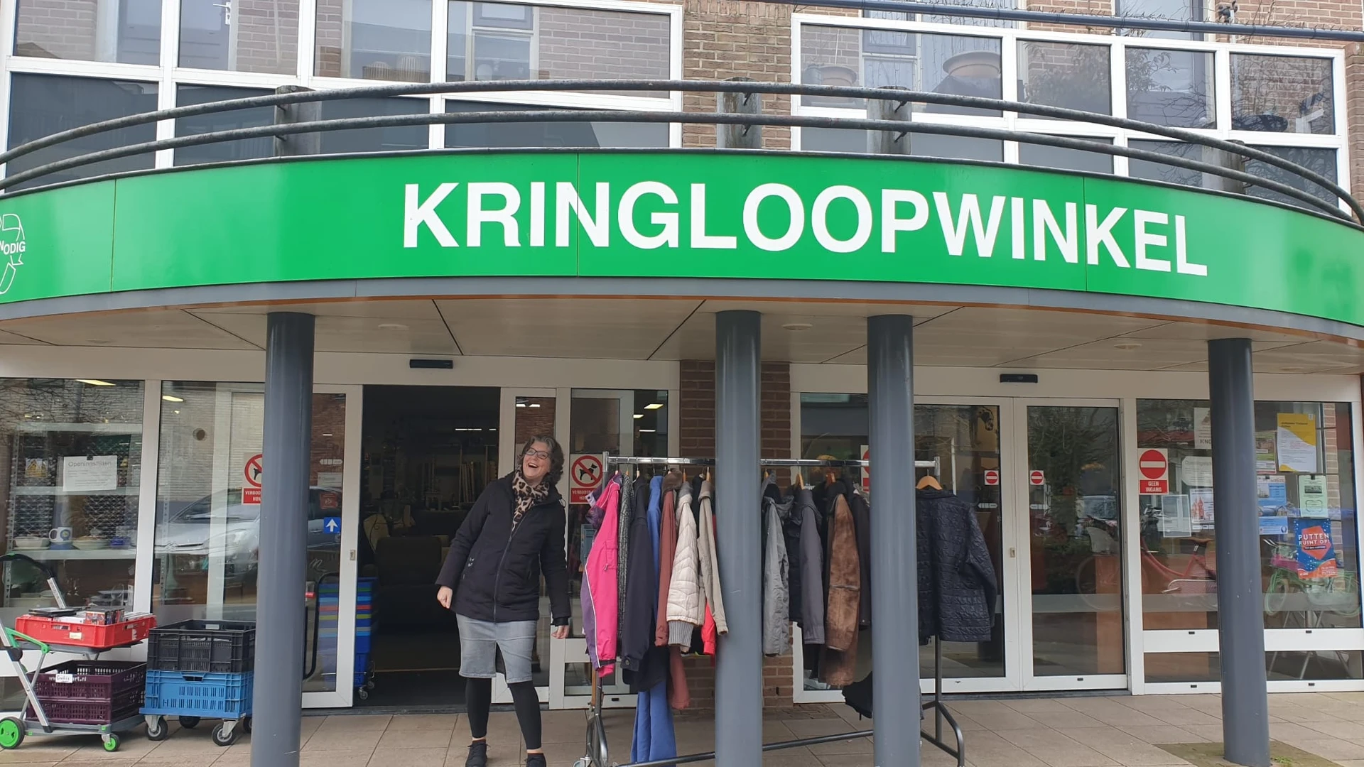 Groene gevel van kringloopwinkel met uitstalraam met kleding, persoon in zwarte jas voor de ingang
