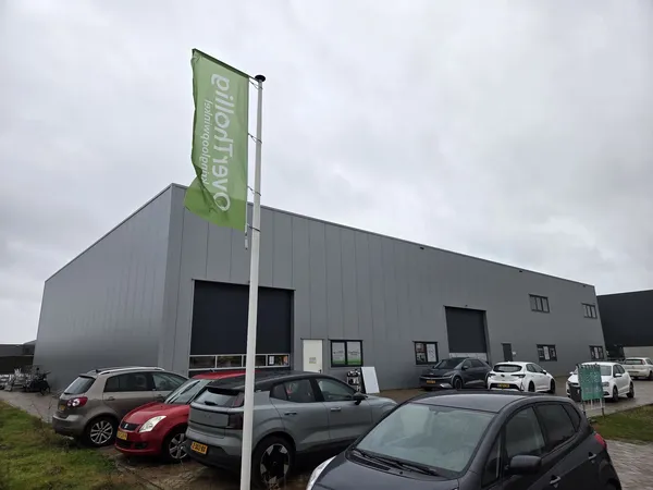 Grijze bedrijfsgebouw met groene vlaggenmaststijl en parkeerplaats met auto's