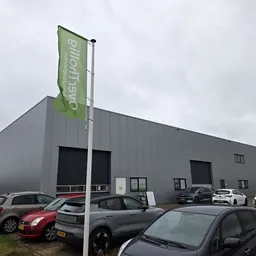 Grijze bedrijfsgebouw met groene vlaggenmaststijl en parkeerplaats met auto's