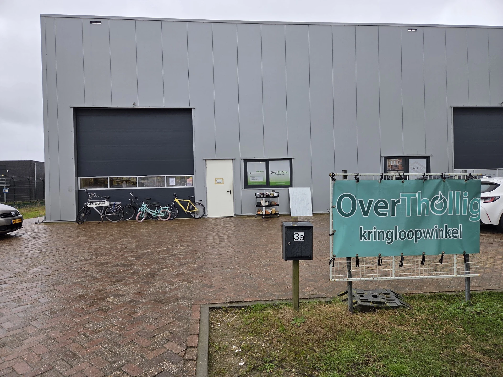 Grijze gevel van kringloopwinkel met twee donkere roldeurenaan voorkant en groen bord met winkelnaam OverThollig