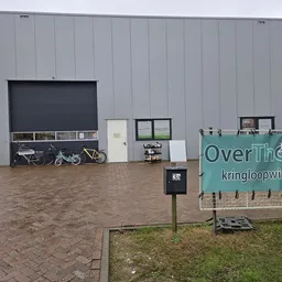 Grijze gevel van kringloopwinkel met twee donkere roldeurenaan voorkant en groen bord met winkelnaam OverThollig
