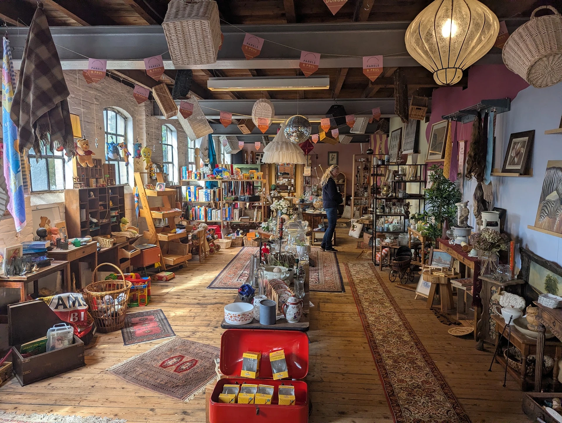 Ruim winkelbinnenaanzicht van kringloopwinkel met houten vloer, stellingen, hangende lampionnen en diverse tweedehands spulle
